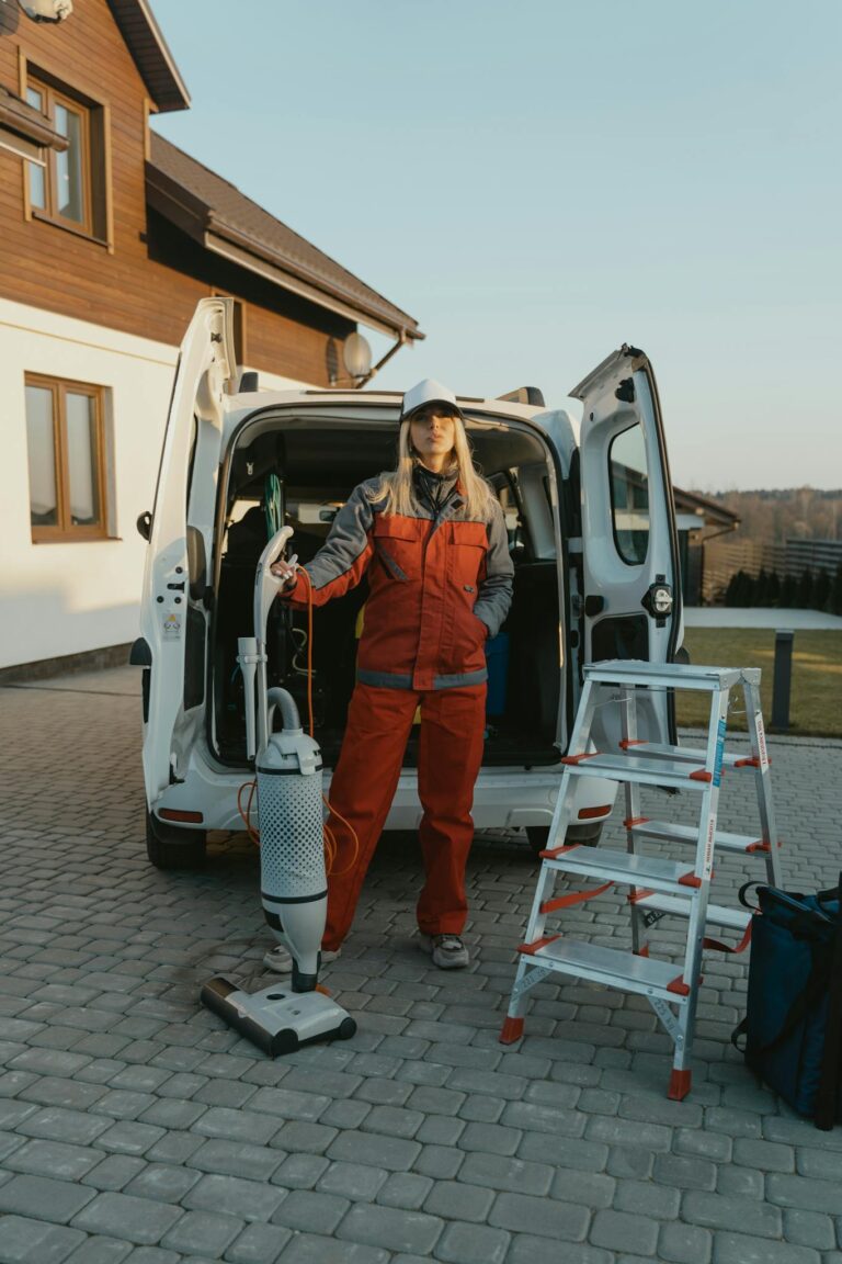 A cleaner in uniform stands beside a van with cleaning tools outside a modern house at sunset.